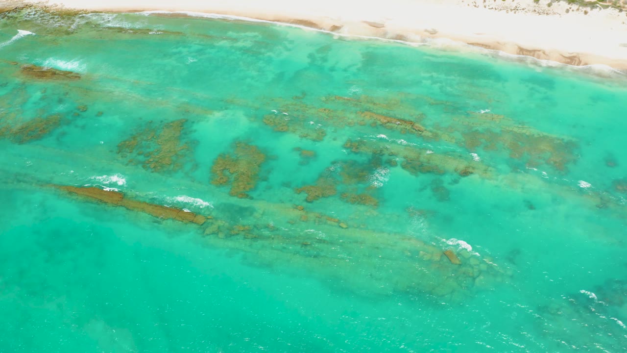 Cabo Pulmo aerial view showing a narrow sandy shore at the top, long brown submerged rock shapes through the middle, small foam streaks, and no visible people.