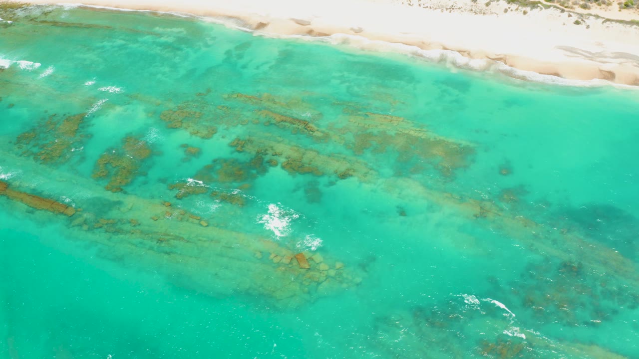 Aerial frame of Cabo Pulmo shallows with coral and rock shapes under turquoise water, a narrow sandy shoreline above, and no visible people.