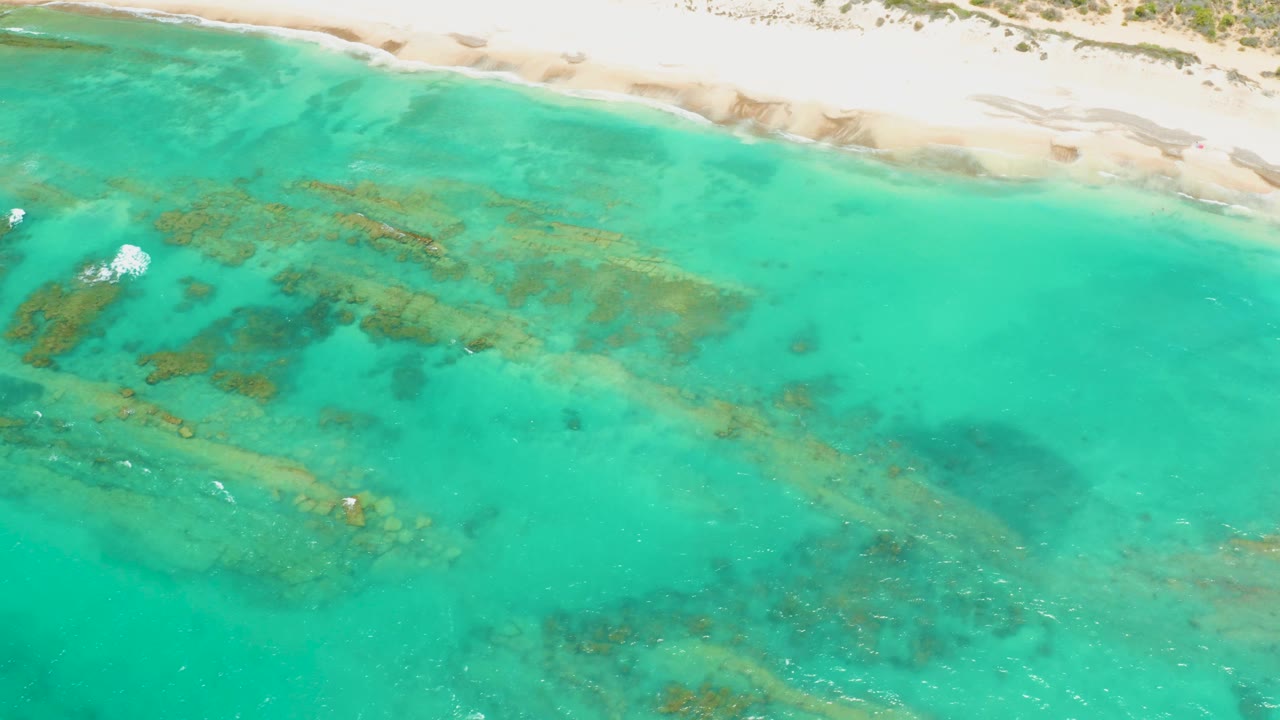 Aerial view over Cabo Pulmo shallow water with darker reef shapes across the center, a narrow sandy beach at the top, small foam at left, and no visible people.