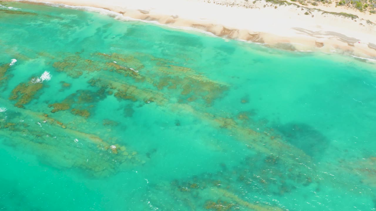 Cabo Pulmo aerial frame with bright turquoise water, dark seabed areas beneath the surface, pale sand across the top, small breaking foam at left, and no visible people.