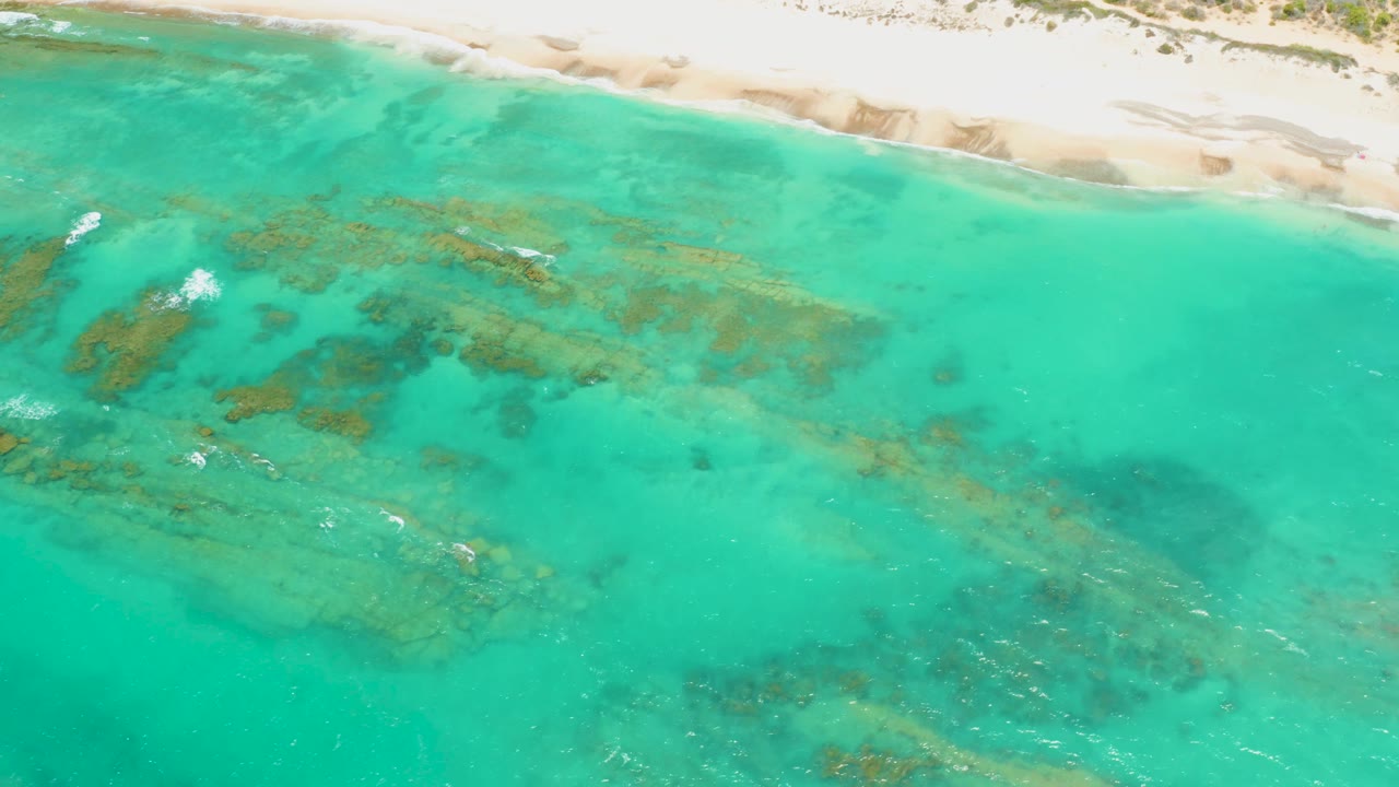 Aerial Cabo Pulmo frame showing shallow turquoise ocean, reef and rock texture running diagonally through the water, pale sand near the top edge, and no visible people.