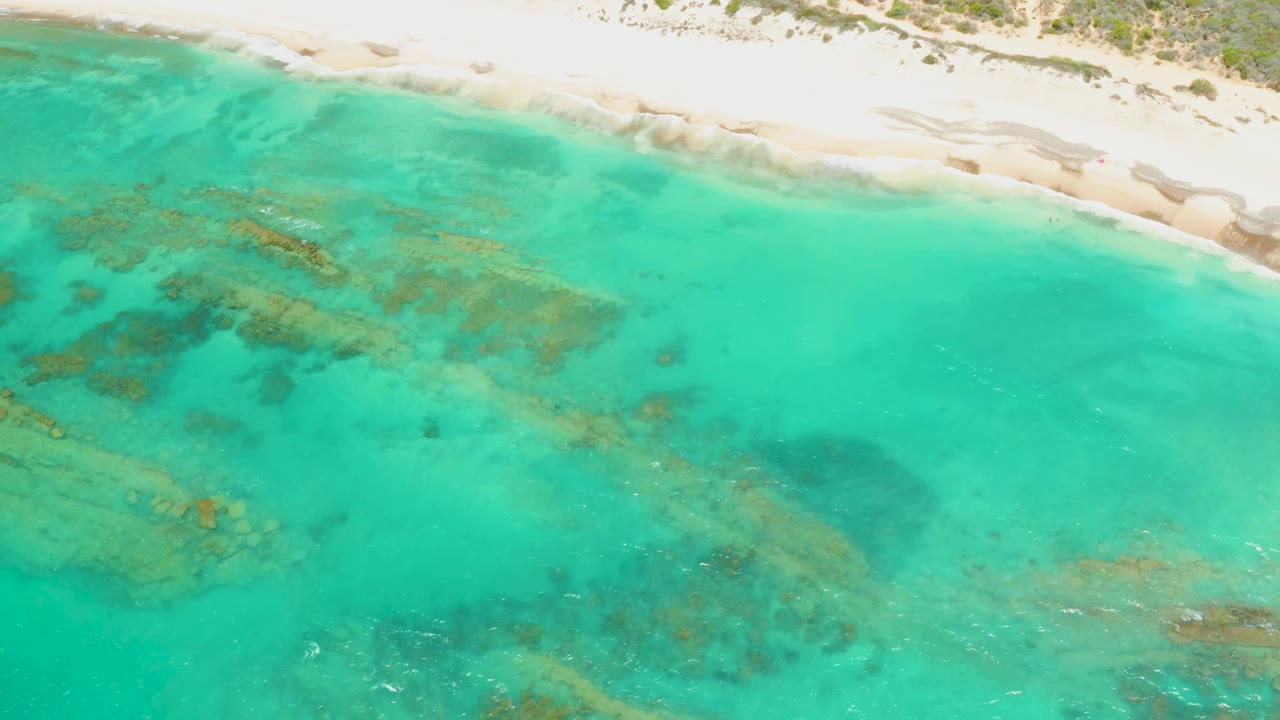 Overhead view of Cabo Pulmo turquoise shallow water with submerged rock patches and a pale sandy shoreline at the top, no visible people.