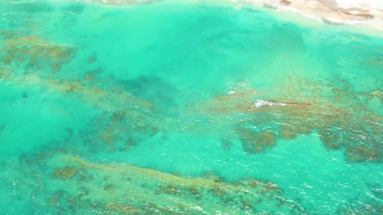 Overhead Cabo Pulmo frame showing clear turquoise water, brown-green reef patches, small white foam near the center-right, and no visible people.