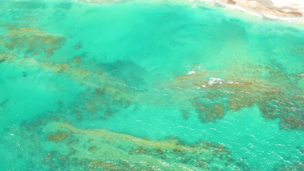 Aerial view in Cabo Pulmo with reef bands under bright turquoise water, a narrow sandy beach at the upper edge, and no visible people.