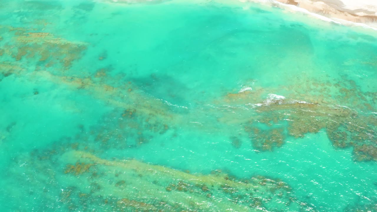 Mid-clip overhead frame of Cabo Pulmo shallows with coral and rock shapes under the water, soft ripples across the surface, and no visible people.