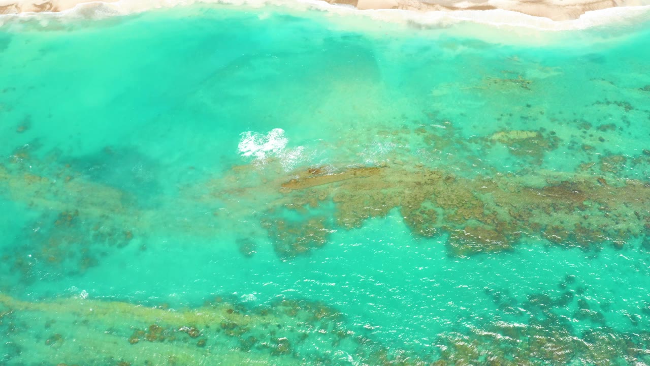 Overhead view of turquoise coastal water at Cabo Pulmo with pale sand at the top edge, darker underwater formations through the shallows, and no visible people.