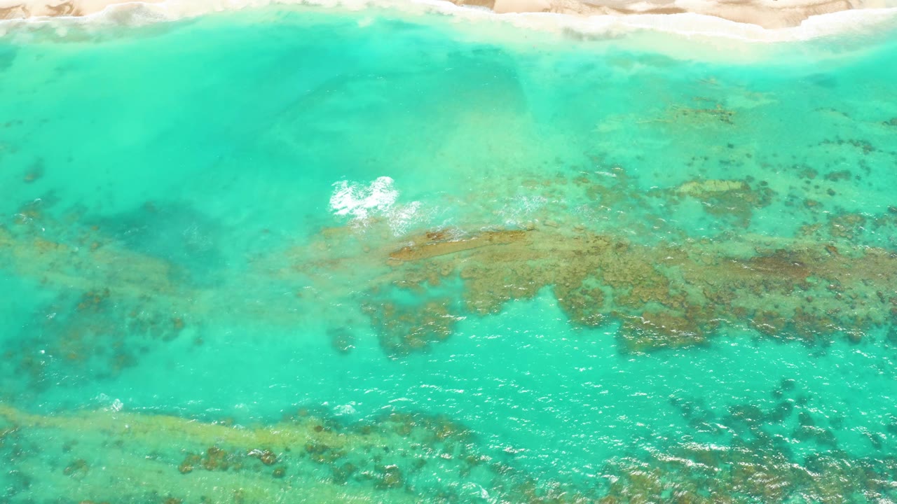 Aerial frame over Cabo Pulmo shallows showing a bright white foam patch near the left center, tan sand above, and reef-like formations visible under turquoise water with no visible people.