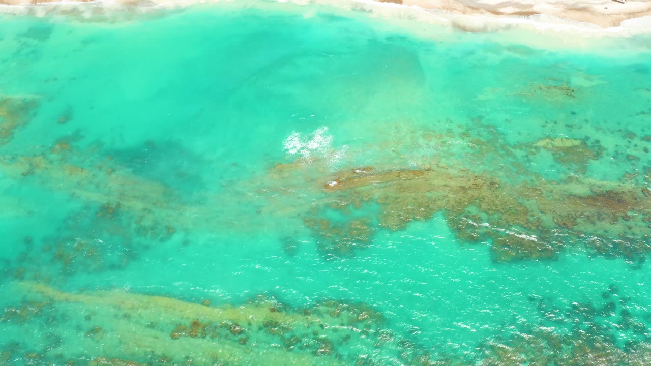 Aerial view of Cabo Pulmo shallows with pale sand running across the upper frame, a small bright foam area left of center, and coral-like shapes below the surface, with no visible people.