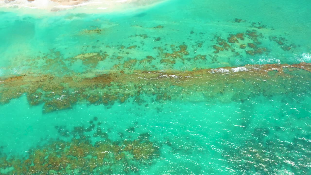 Aerial frame over Cabo Pulmo shallows with scattered dark reef patches below the beach edge, a thin foam streak across the reef, and no visible people.
