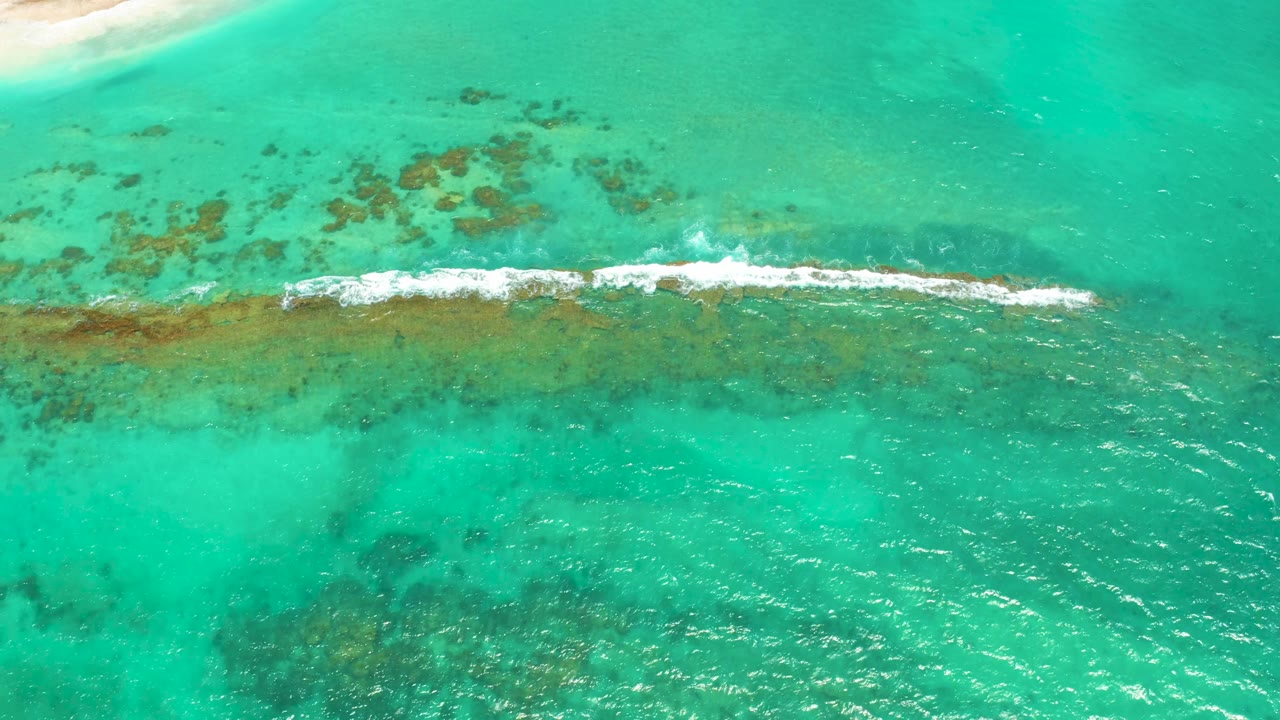 Aerial frame over Cabo Pulmo showing the white surf line stretched across the central reef, with scattered darker reef patches above and no visible people.
