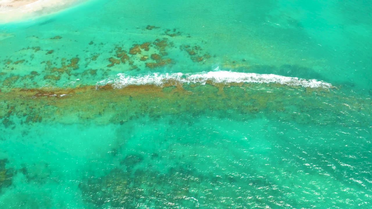 Aerial view of Cabo Pulmo water where white foam thickens along the central reef, darker reef marks spread above it, and no visible people appear.