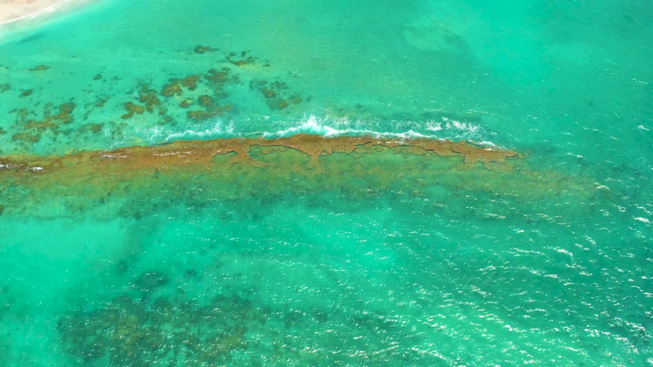 A shallow coral reef crosses the center of the Cabo Pulmo frame as white surf spreads farther along the reef, with darker submerged patches below and no visible people.