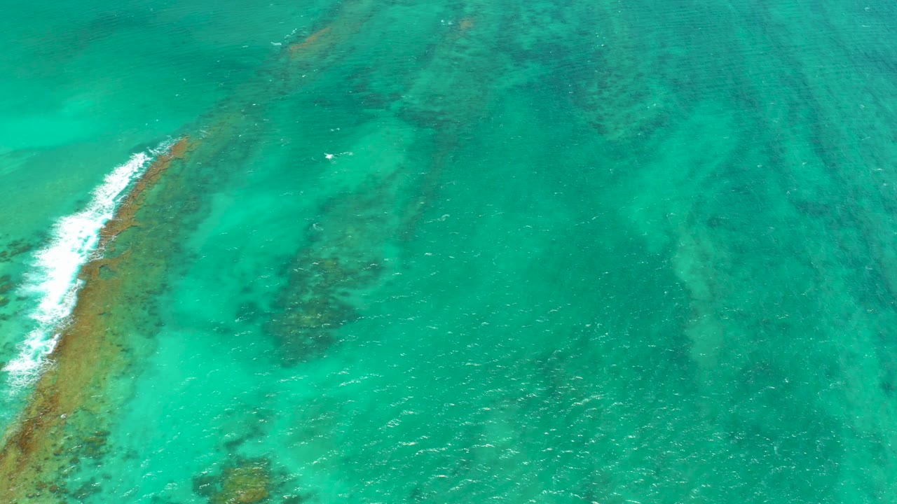 Aerial frame from Cabo Pulmo with a diagonal reef edge, white foam spreading along it, clear green-blue water, and no visible people.