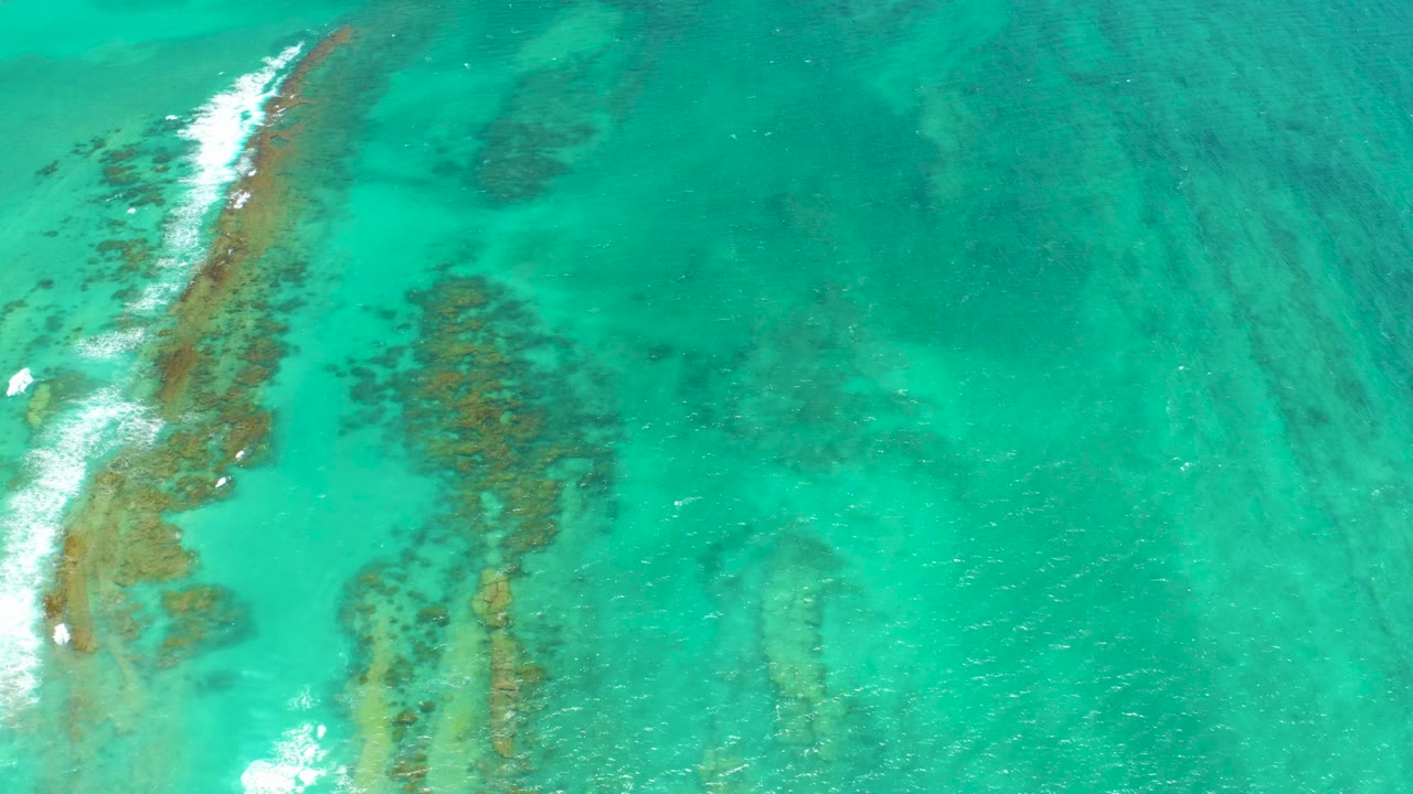 Aerial Cabo Pulmo frame showing clear green-blue water, darker coral formations below the surface, foam continuing down the left reef line, and no visible people.
