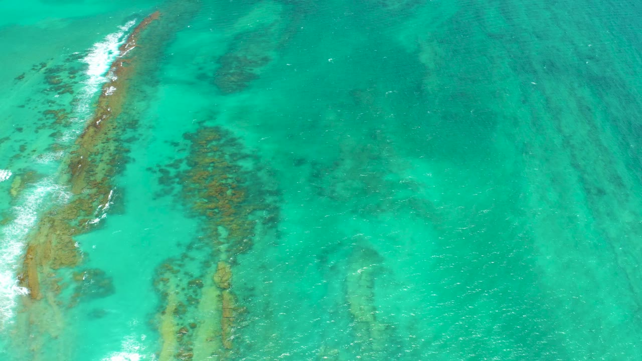 Aerial Cabo Pulmo ocean frame with the reef line close to the left border, pale foam at the lower left, scattered submerged formations, and no visible people.