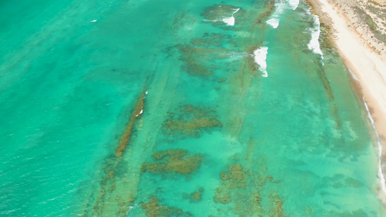 Overhead view of turquoise water and sandy shoreline in Cabo Pulmo, Baja California Sur, with darker underwater formations visible and no visible people.
