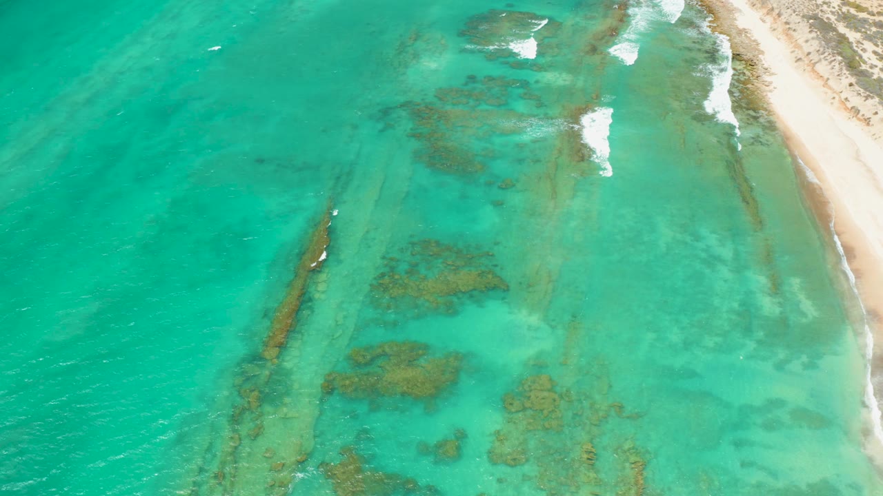 Aerial frame of Cabo Pulmo shoreline with turquoise shallows, a pale sand strip on the right, scattered white foam, and no visible people.