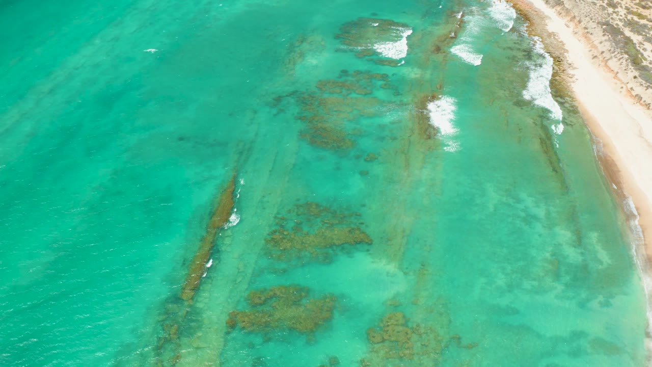 Turquoise coastal water with a long dark underwater formation and sandy beach edge.