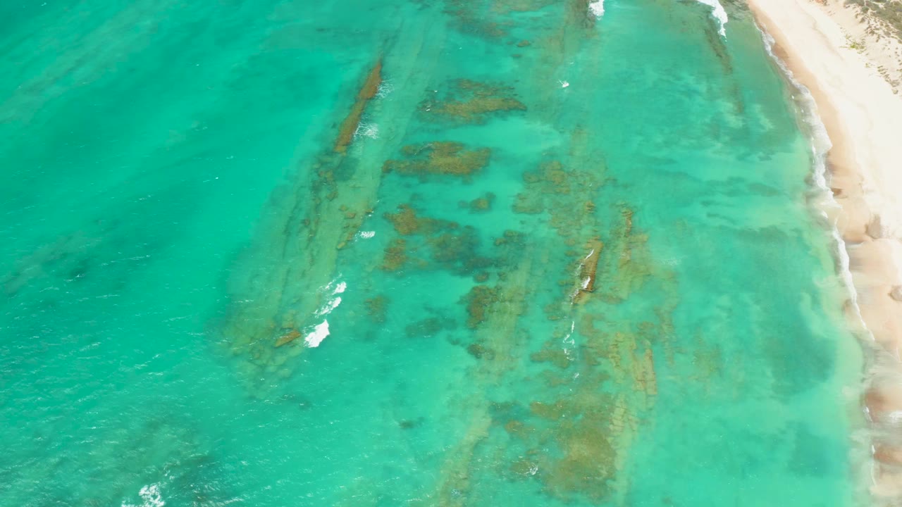 Aerial frame from Cabo Pulmo with darker underwater formations centered beneath clear turquoise water, small white surf marks, beach at right, and no visible people.
