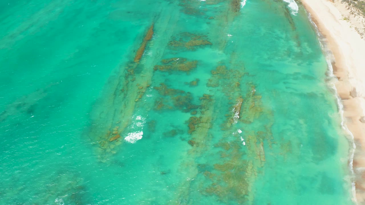 Cabo Pulmo aerial view showing clear water over clustered seabed formations, small white surf breaking near the lower left, beach at right, and no visible people.