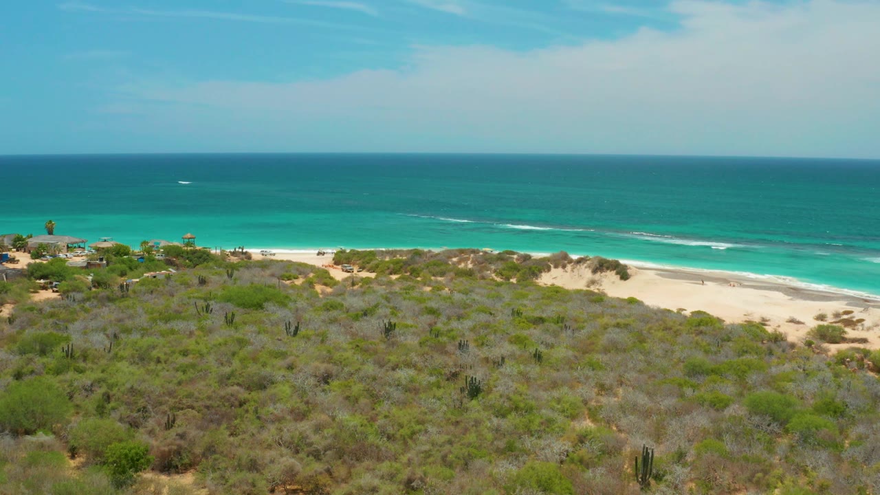 Cabo Pulmo aerial frame showing more of the dune ridge and shoreline, with green scrub in the foreground, blue-green ocean behind it, thin surf lines, and no visible people.
