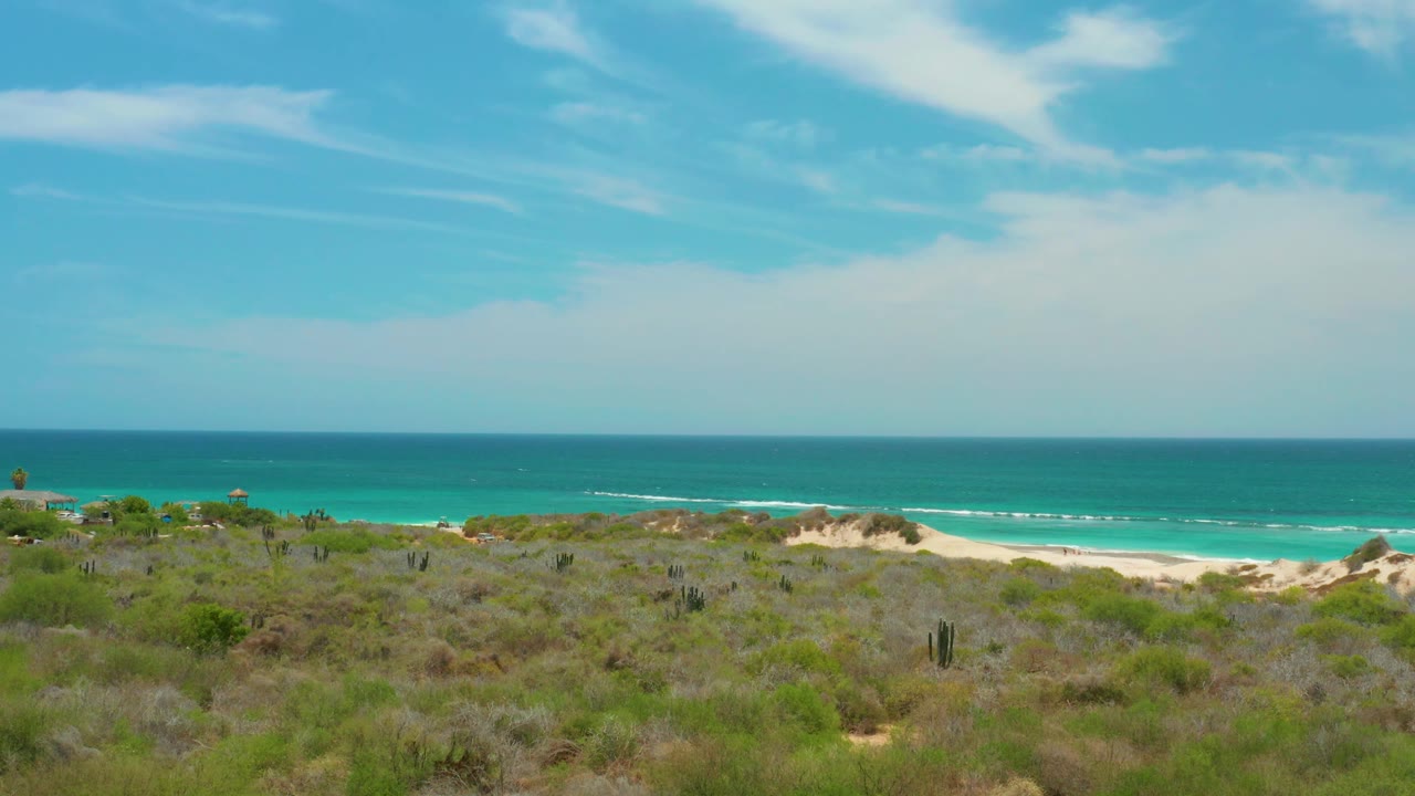 Cabo Pulmo dunes and low vegetation frame a pale beach and blue-green ocean, with a broad cloud band across the sky and no visible people.