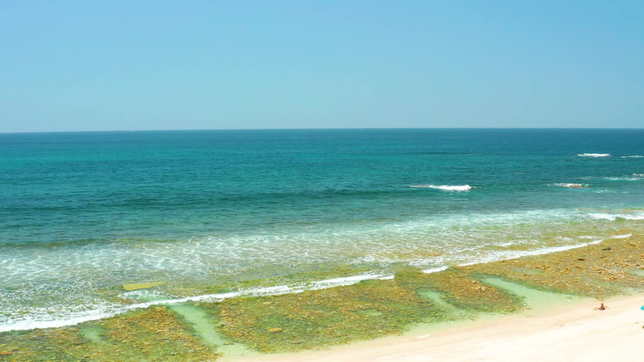 Aerial view of El Tule Beach in Los Cabos with pale sand at the bottom, turquoise shallows over reef patches, blue ocean beyond, and no visible people.