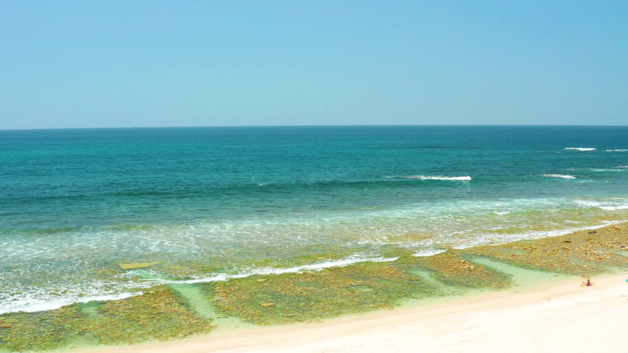 Aerial frame over El Tule Beach showing pale sand across the lower edge, reef-textured shallow water, white wave breaks, blue ocean, and a tiny distant beachgoer near the right side.