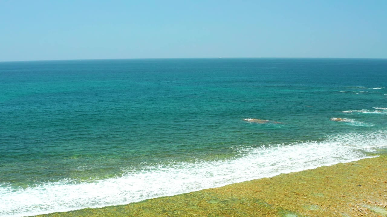 El Tule Beach Los Cabos shoreline with a diagonal band of white foam pushing across clear turquoise water toward the sandy lower edge, with no visible people.