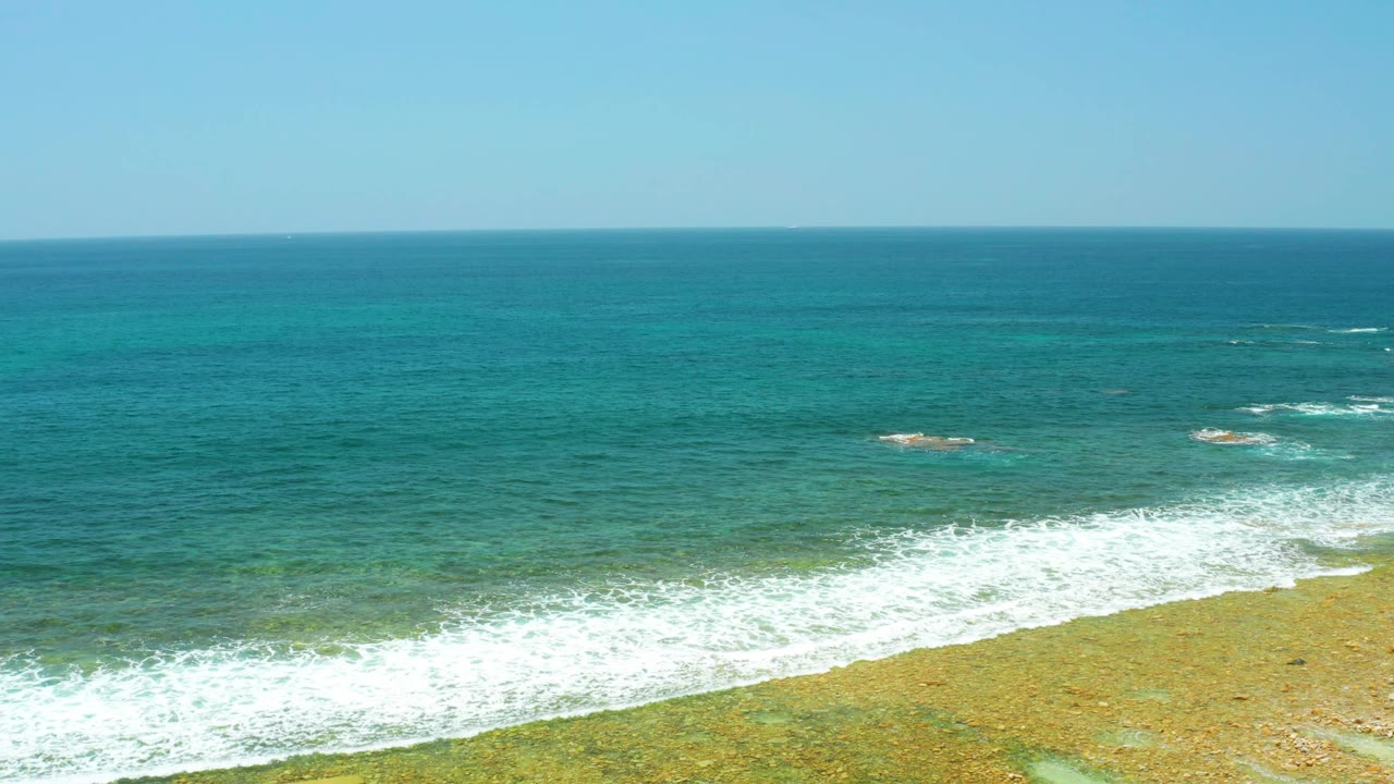 Wide elevated frame at El Tule Beach Los Cabos showing teal ocean under clear sky, white breakers near shore, and exposed sandy shallows with no visible people.