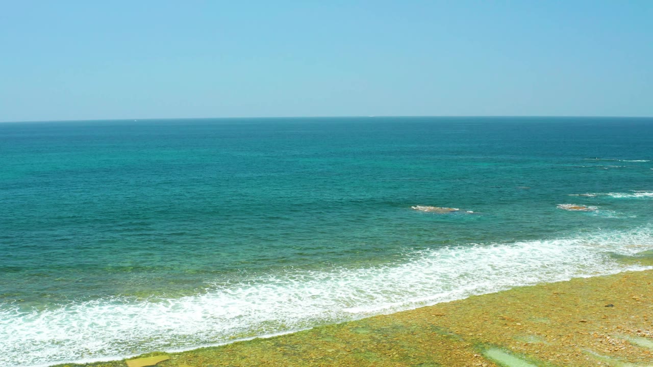 Aerial-style view of El Tule Beach Los Cabos with bright turquoise sea, white wave foam across the lower frame, shallow reef texture, and no visible people.