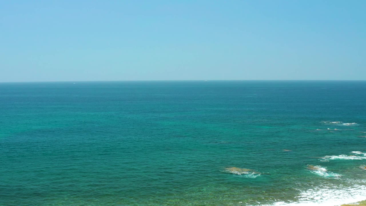 Aerial-looking view at El Tule Beach with clear sky, blue-green ocean, foamy white surf beginning to show near the lower right edge, and no visible people.