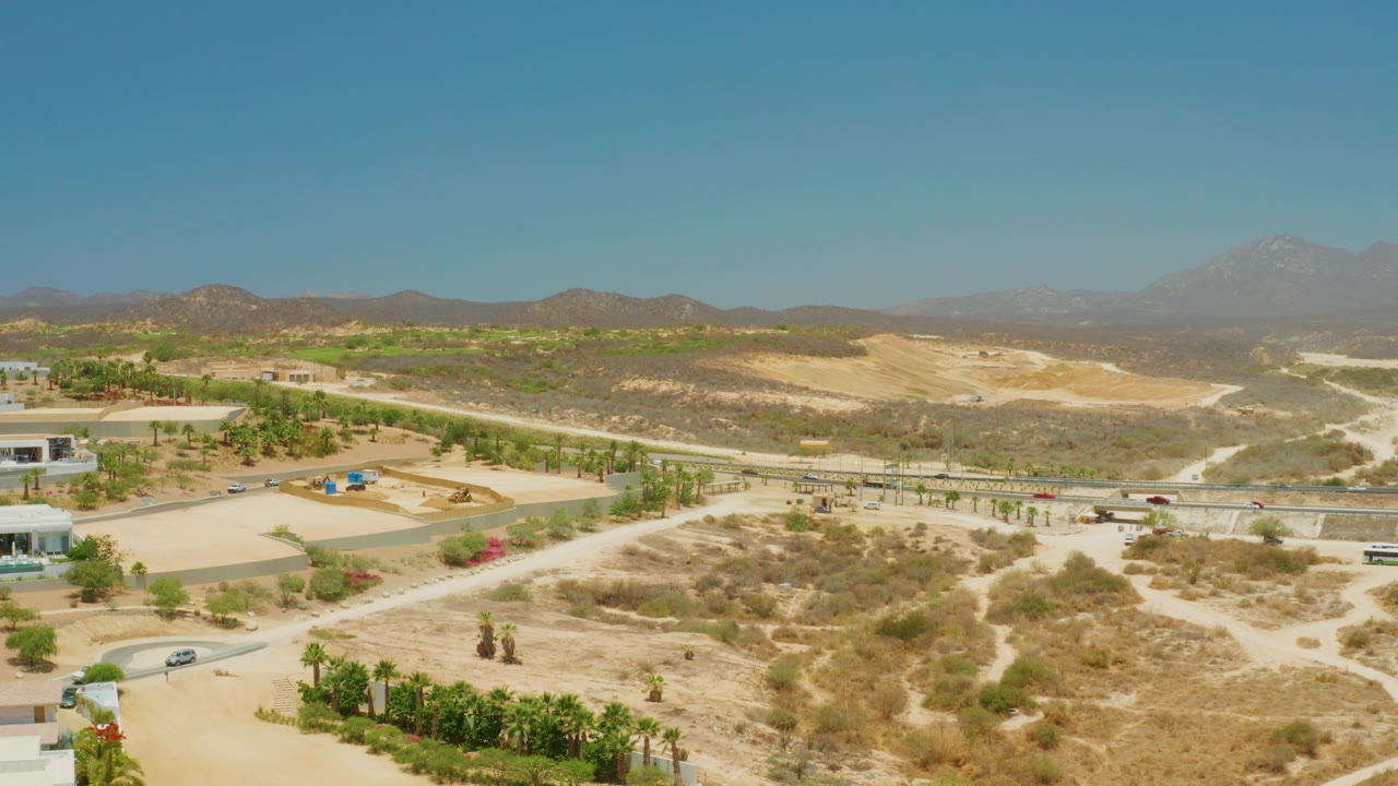 Aerial view at 0:01 over El Tule Beach Los Cabos with vehicles on a midground road, sandy washes, sparse desert plants, palm rows, distant ridges, and no visible people.