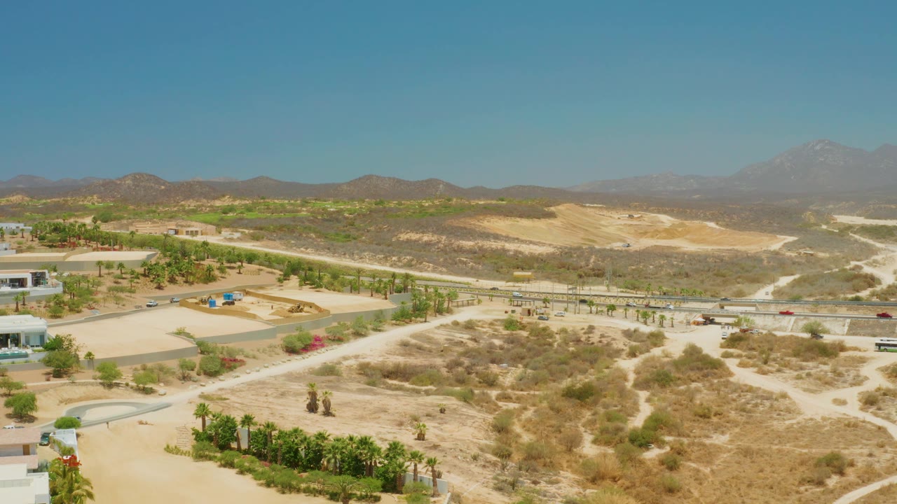 Aerial frame from El Tule Beach Los Cabos showing roadside vehicles, pale dirt tracks, planted palms, dry brush, low development, hazy mountains, and no visible people.