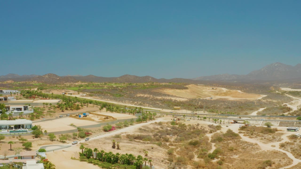 At 0:00, El Tule Beach Los Cabos is seen from above with sandy roads, scattered shrubs, low buildings, small vehicles, distant mountains, and no visible people.
