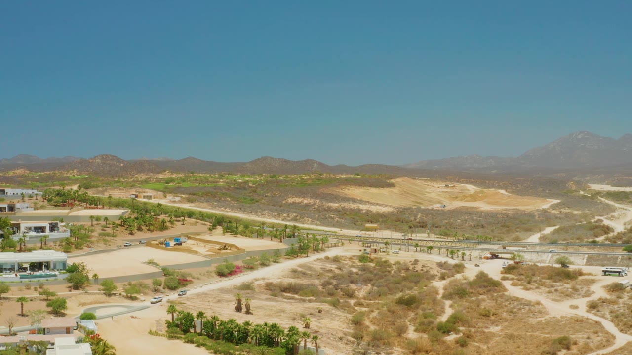 At 0:01, the aerial view over El Tule Beach Los Cabos holds on dry road corridors, patches of greenery, low structures, vehicles, hazy mountains, and no visible people.