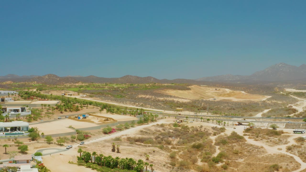 At 0:02, El Tule Beach Los Cabos appears as an arid expanse with a visible road line, small cars, planted greenery, sandy slopes, distant mountains, and no visible people.