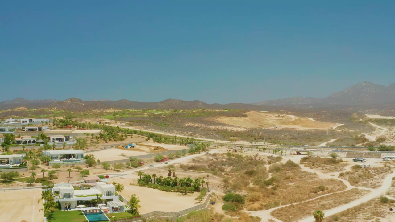 Opening aerial view over El Tule Beach Los Cabos with modern villas, palms, pale dirt tracks, dry hills, distant mountains, and no visible people.