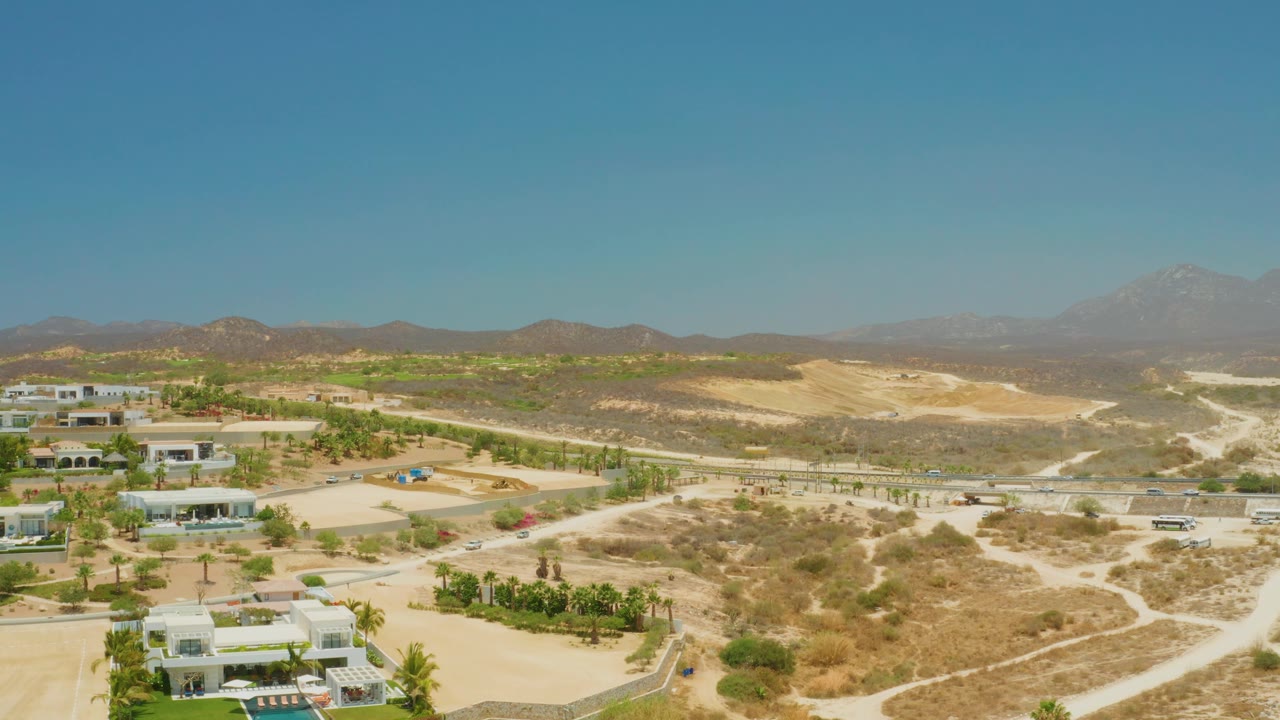 Wide aerial view over El Tule Beach Los Cabos with white villas at left, scrub-covered terrain across the center, hazy mountains, and no visible people.