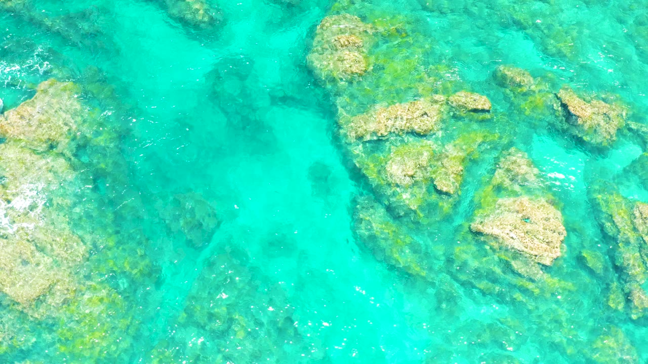 Aerial frame over El Tule Beach water where clustered submerged rocks sit near the upper right and darker seabed patches show through the turquoise surface, with no visible people.
