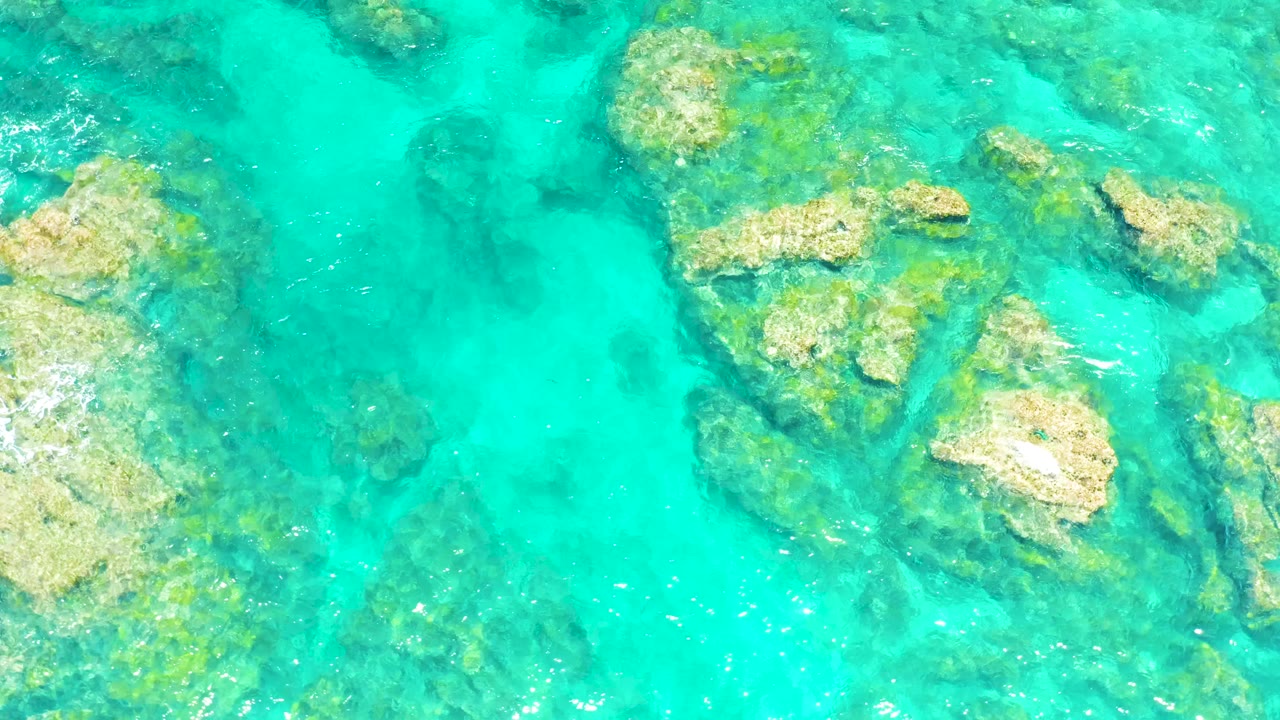 Overhead El Tule Beach frame with bright turquoise water crossing algae-tinted rock ledges and a larger pale rock near the lower right, with no visible people.