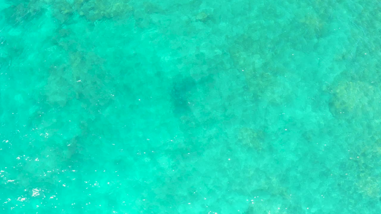 Top-down view of turquoise ocean water at El Tule Beach, Los Cabos, with pale seabed texture and small sun glints, no visible people.
