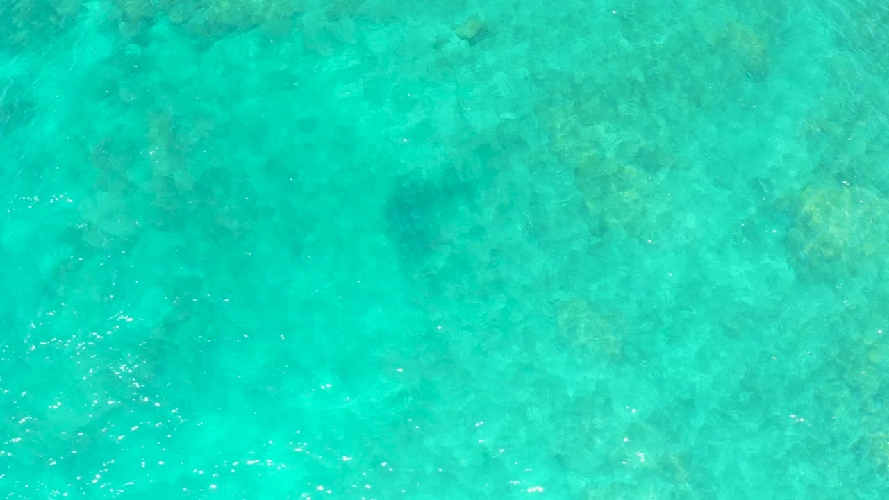 Top-down El Tule Beach water view with blue-green surface color, scattered darker seabed patches, and bright specks of reflected sunlight, no visible people.