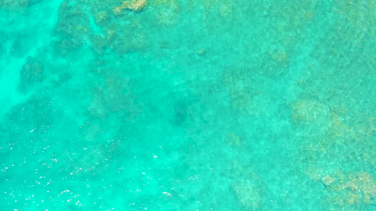 Overhead view of clear turquoise water at El Tule Beach Los Cabos with darker submerged rock patches near the top edge and no visible people.