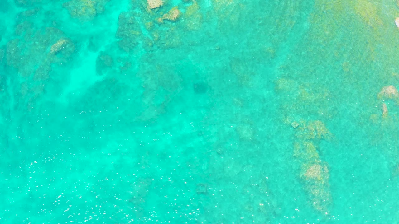 Top-down view of shallow turquoise ocean at El Tule Beach Los Cabos with reef-like rocks beneath the surface and bright specks across the water, no visible people.