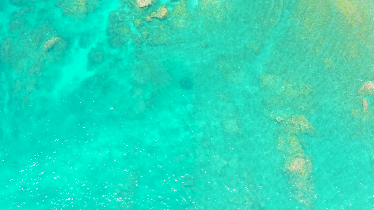 Overhead frame from El Tule Beach Los Cabos showing turquoise shallow water, a rocky seabed, and soft surface texture with no visible people.