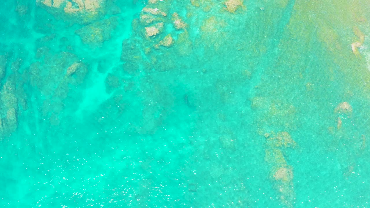 Top-down view of turquoise shallow water at El Tule Beach in Los Cabos, with submerged rock patches near the upper edge and no visible people.