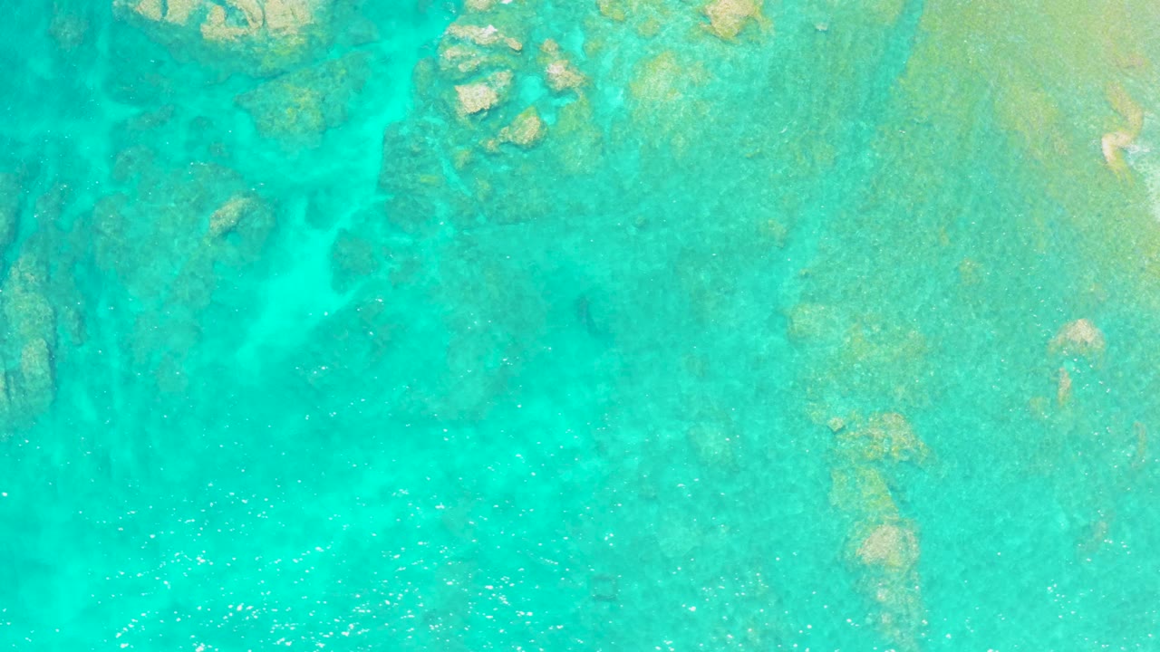 Clear turquoise water at El Tule Beach in Los Cabos shows a rocky seabed through the surface, with small white sparkles across the lower portion and no visible people.