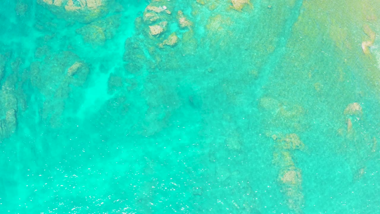Blue-green shallow ocean water at El Tule Beach in Los Cabos fills the frame, with pale submerged rocks on the right and no visible people.