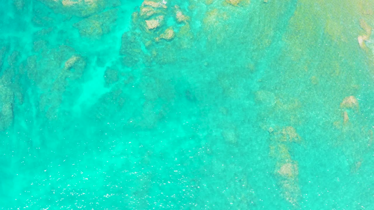 Turquoise shallow water with darker rock shapes and bright surface flecks.